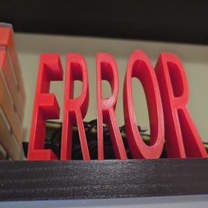 以下が含まれることがあります： 「ERROR」という単語が書かれた赤い3Dプリントのサイン。文字は太く、暗い面に立っています。サインの左側には、オレンジと透明なプラスチック容器のスタックが見えます。