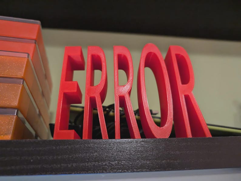 以下が含まれることがあります： 「ERROR」という単語が書かれた赤い3Dプリントのサイン。文字は太く、暗い面に立っています。サインの左側には、オレンジと透明なプラスチック容器のスタックが見えます。