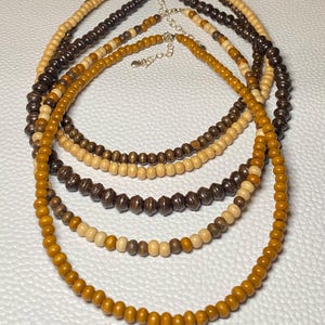May include: Five wooden bead necklaces in varying shades of brown and tan. The necklaces have silver-colored clasps and are arranged in a layered display. The beads are round and smooth, creating a natural and earthy aesthetic.