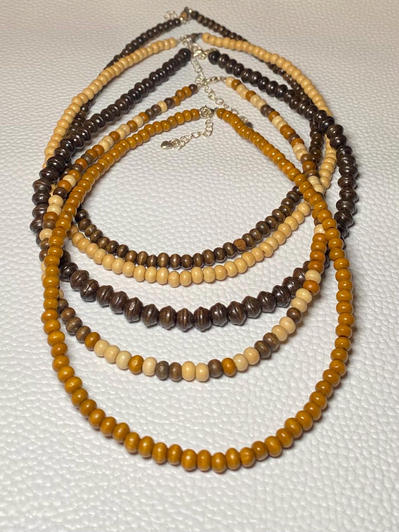 May include: Five wooden bead necklaces in varying shades of brown and tan. The necklaces have silver-colored clasps and are arranged in a layered display. The beads are round and smooth, creating a natural and earthy aesthetic.