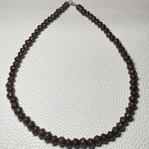 May include: A dark brown beaded necklace. The beads are small, cylindrical, and evenly spaced. The necklace has a silver-colored chain and clasp. The beads appear to be made of wood or a similar material. The necklace is displayed on a white surface.
