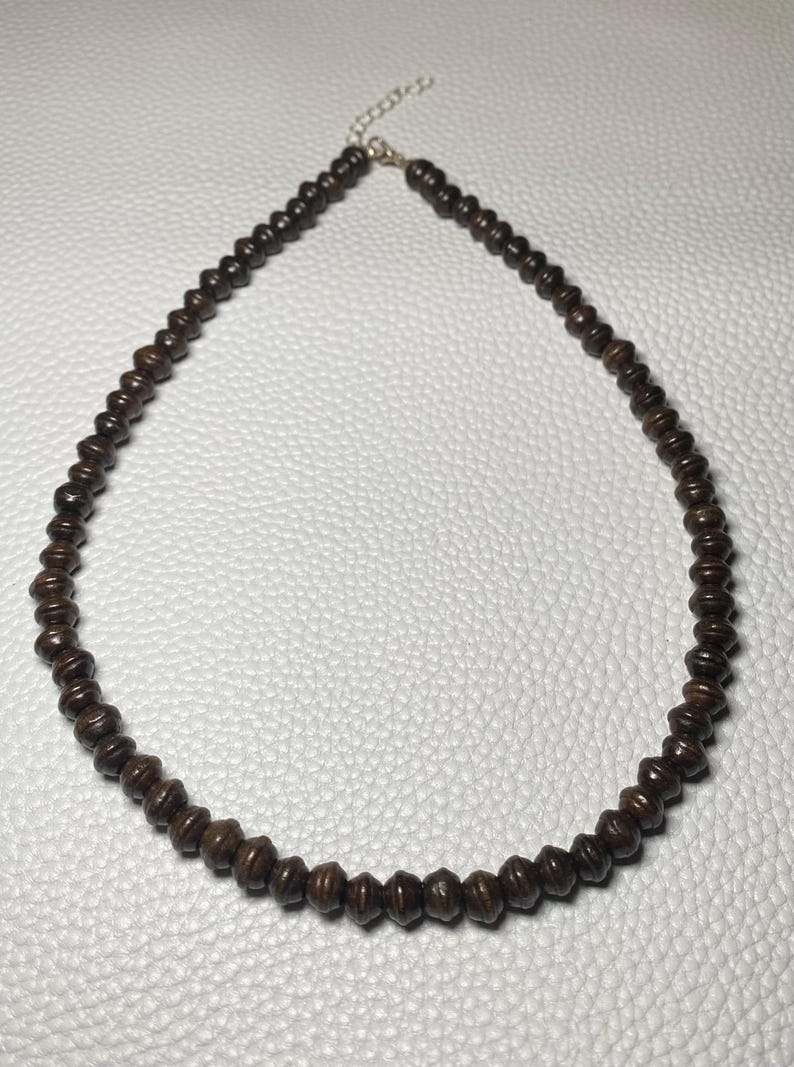 May include: A dark brown beaded necklace. The beads are small, cylindrical, and evenly spaced. The necklace has a silver-colored chain and clasp. The beads appear to be made of wood or a similar material. The necklace is displayed on a white surface.