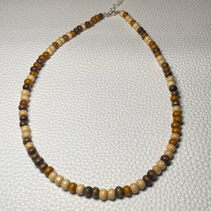 May include: A beaded necklace featuring a variety of wooden beads in shades of brown and tan. The necklace has a silver-colored clasp and is designed to be worn around the neck. The beads are uniform in size and shape.