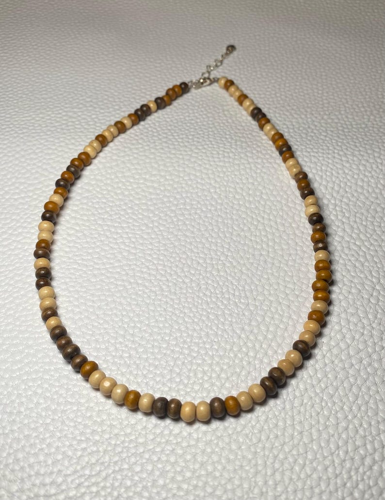 May include: A beaded necklace featuring a variety of wooden beads in shades of brown and tan. The necklace has a silver-colored clasp and is designed to be worn around the neck. The beads are uniform in size and shape.