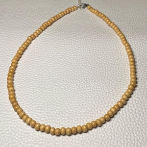 May include: A light brown beaded necklace. The necklace is made of small, round wooden beads strung together. The necklace has a silver-colored clasp and is displayed on a white surface.