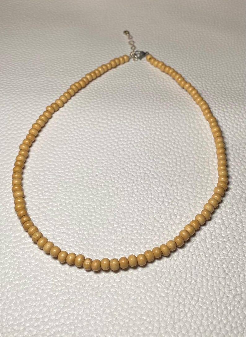 May include: A light brown beaded necklace. The necklace is made of small, round wooden beads strung together. The necklace has a silver-colored clasp and is displayed on a white surface.