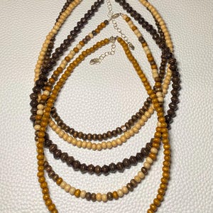 May include: A collection of six wooden bead necklaces in varying shades of brown and tan. The necklaces have different bead shapes and sizes, with adjustable silver-tone clasps. The beads range from light beige to dark brown.