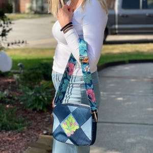 May include: A denim crossbody bag with a floral strap. The bag features a patchwork design with a central embroidered floral motif in pink and green. The strap is navy blue with pink and white flowers.
