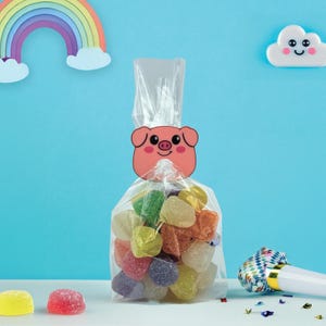 May include: A clear cellophane bag filled with colorful gummy candies, topped with a pink pig-shaped tag. The background features a rainbow and a smiling cloud against a blue backdrop. Party favors.