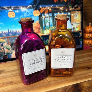 May include: Two decorative glass bottles with cork stoppers. One is purple and the other is orange. Each bottle has a white label with black text that reads "SALLY'S DEADLY LIGHT SHADE" and "SALLY'S WORMWORT" respectively. The bottles are filled with wax melts.