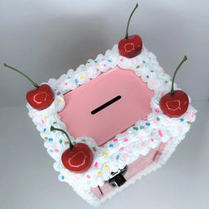May include: A pink metal box with a slot, decorated with white frosting, colourful sprinkles, and four red cherries. The box has a small lock and is designed to look like a dessert.