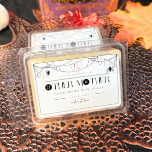 Closeup of front label on two Other Mother wax melts, showing black syrup swirl and black button accents on white square soy wax, Coraline inspired spooky Halloween fragrance.