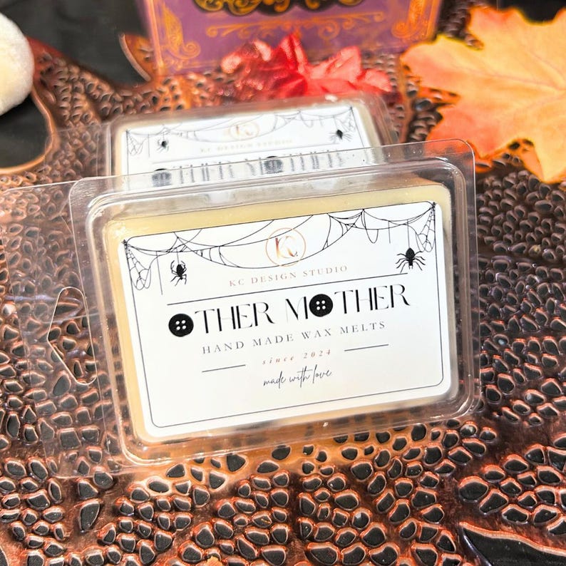 Closeup of front label on two Other Mother wax melts, showing black syrup swirl and black button accents on white square soy wax, Coraline inspired spooky Halloween fragrance.