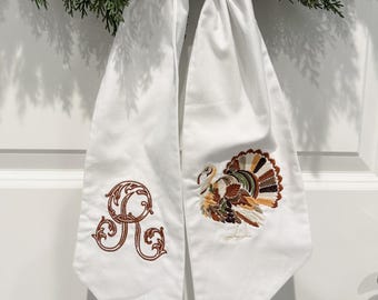 May include: White fabric Thanksgiving decoration with an embroidered turkey and the letter "R". The turkey has brown, green, and orange feathers. The decoration is tied to a green wreath.