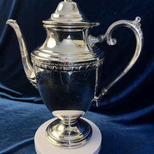 May include: A polished silver coffee pot featuring a tall, slender body, a curved spout, and an ornate handle. The lid is topped with a decorative finial, and the midsection is adorned with a detailed embossed band. It stands on a circular base.