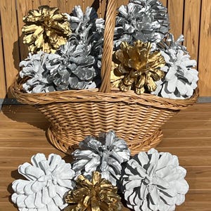 May include: A woven wicker basket with a central handle filled with multiple pinecones painted in metallic gold, silver, and matte white. Three additional painted pinecones in gold, silver, and white are arranged in front of the basket.