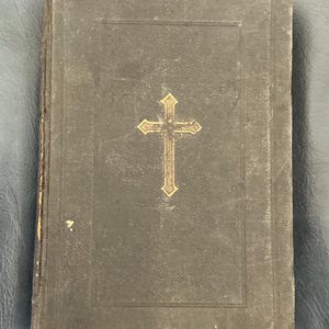 May include: A dark gray, antique book with a gold cross emblem on the cover. The book's cover has a textured appearance and shows signs of wear, suggesting its age. The cross is centered and has decorative detailing.