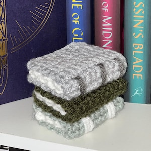 May include: A stack of three crocheted washcloths in shades of gray, olive green, and white. The washcloths are stacked on a white shelf in front of a row of books. The books have titles like "Throne of Glass" and "The Assassin's Blade."