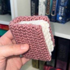 May include: A crocheted book sleeve, resembling a sandwich. The exterior is a dusty rose colour, with a white interior. The sleeve is designed to protect books. The item is handmade and visible against a backdrop of bookshelves.