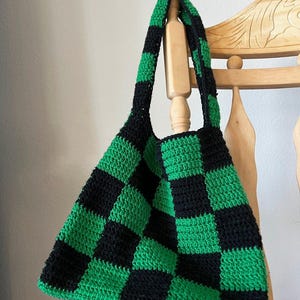 Può includere: Una borsa tote all'uncinetto fatta a mano con un sorprendente motivo a scacchiera in bianco e nero. La borsa è dotata di un ampio e comodo cinturino ed è appesa a un attaccapanni in legno. Il design della borsa è audace e accattivante.