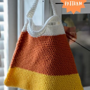May include: A handmade, knitted tote bag in the shape of a candy corn. The bag features a white top, an orange middle, and a yellow bottom. The bag has a white knitted strap and the text "PDF PATTERN" in an orange starburst.