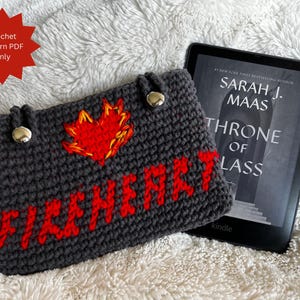 Könnte beinhalten: Eine dunkelgraue gehäkelte Kindle-Hülle mit dem Wort "FIREHEART" in roten Buchstaben und einem roten und gelben Flammendesign. Die Hülle hat zwei silberne Knopfverschlüsse. Ein Kindle-E-Reader mit dem Buch "Throne of Glass" liegt neben der Hülle.