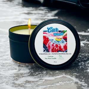 May include: A lit soy wax candle in a black tin with a yellow interior. The lid features a graphic of a red car being washed, with the text "Car Wash". The candle is 8 oz.