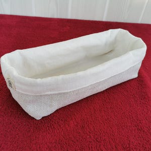 Könnte beinhalten: Ein rechteckiger Brotkorb mit weißem Leineninnenraum und einer strukturierten, hellfarbigen Außenseite. Der Korb steht auf einer roten Oberfläche, und das Innenfutter ist über den Rand gefaltet.