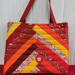 Peut inclure: Sac fourre-tout matelassé avec un motif patchwork aux tons de rouge, orange et jaune. Le sac présente un motif en chevrons avec des accents de tissu floral et à pois. Il a une sangle rouge unie et une petite étiquette circulaire.