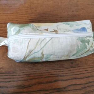May include: A rectangular pouch with a floral pattern, a white zip, and a small fabric loop at each end. The pouch has a cream background with green, blue, and brown floral designs. The zip is white, and the pouch is on a wooden surface.