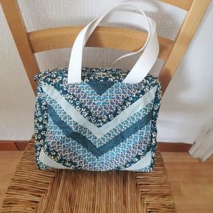Puede incluir: Bolso tote cuadrado con asas blancas, sobre una silla de madera. El bolso presenta un diseño patchwork con fondo verde azulado y estampados florales y geométricos en azul y blanco.