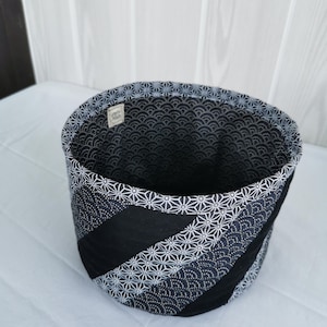 May include: A round fabric storage basket with a black and white geometric pattern. The exterior features diagonal stripes of solid black, dotted, and starburst designs. The interior is lined with a wave pattern.