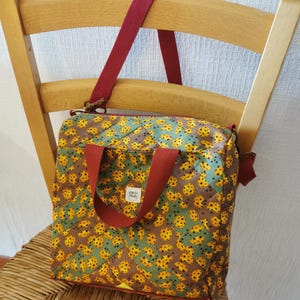 Peut inclure: Sac fourre-tout en tissu matelassé avec un motif répété de formes jaunes et marron sur un fond vert et marron. Le sac a une poignée et une bandoulière rouges, posé sur une chaise en bois.