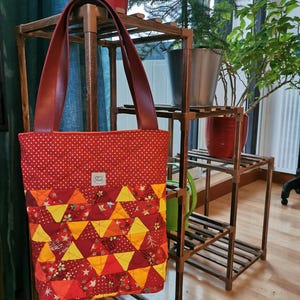 Peut inclure: Un sac fourre-tout rouge avec des poignées bordeaux. Le sac présente un motif patchwork avec des pois rouges et un motif géométrique de triangles jaunes, orange et rouges. Le sac est présenté sur un support à plantes en bois.