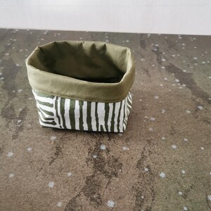 以下が含まれることがあります： 緑色の内側と縁が付いた、小さな布製の収納バスケット。外側は白と緑のストライプ模様です。バスケットは直立するように設計されており、小さなアイテムを整理するために使用される可能性があります。