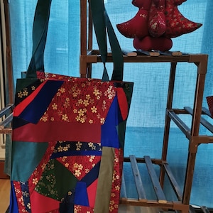 Peut inclure: Un sac fourre-tout patchwork avec une poignée verte et un design coloré. Le sac présente du tissu rouge, bleu et vert avec un motif floral. Une décoration de sapin de Noël rouge est posée sur une étagère en bois.