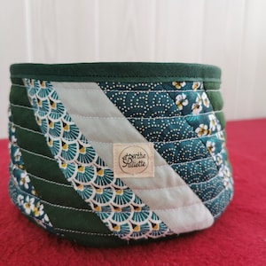 Peut inclure: Un panier en tissu matelassé avec un bord vert foncé et un motif patchwork. Le patchwork comprend des motifs de fleurs blanches, de pois turquoise et un motif en forme d'éventail turquoise et jaune. Une petite étiquette avec le texte "Berthe & Juliette" est cousue sur le côté.