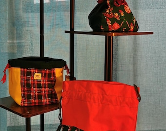 Pode incluir: Um expositor de vários níveis contém várias bolsas com cordão. As bolsas apresentam vários padrões, incluindo um design floral preto, um estampado xadrez vermelho e preto e uma estampa verde com tema natalino com elfos e ornamentos.