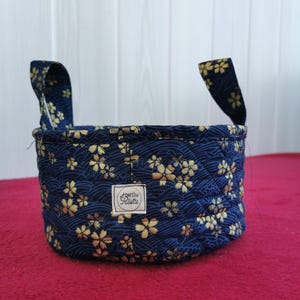 Peut inclure: Un petit panier rond en tissu avec deux poignées. Le panier est bleu marine avec un motif répété de fleurs dorées. L'étiquette sur le devant indique "perthe Palette".