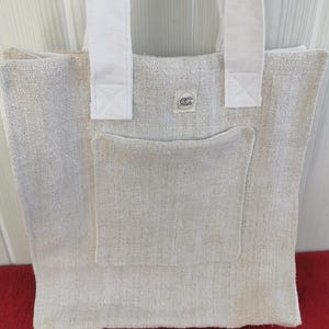 May include: A beige tote bag made of a coarse, woven fabric featuring two white shoulder straps and a large square front pocket. A small rectangular label with the text Bertha Juliette is stitched onto the upper center of the bag body.