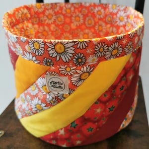 Peut inclure: Un panier rond en tissu avec un motif patchwork. L'intérieur et le bord supérieur présentent un motif floral de marguerites sur fond orange. L'extérieur présente des rayures diagonales de jaune, de rouge avec des étoiles et de bordeaux. Une petite étiquette indique "Berthe Juliette".