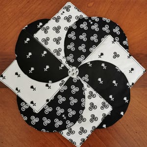 Peut inclure: Un tapis en tissu en forme de moulinet avec un motif répétitif noir et blanc. Le design alterne entre des sections de tissu blanc à petits motifs floraux noirs et de tissu noir à symboles triskèles blancs, assemblées au centre.