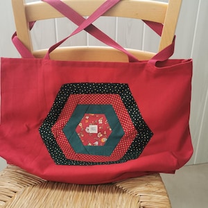 Peut inclure: Un sac fourre-tout rouge avec un motif hexagonal en patchwork. Le sac a des sangles rouges et un hexagone décoratif au centre, avec des tissus verts, rouges à pois et noirs à pois. Le sac est posé sur une chaise en bois.