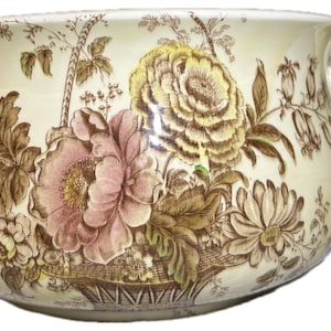 May include: A cream-colored ceramic chamber pot featuring a brown and pink floral transferware pattern. The vessel has a wide, rounded body, a flared rim, and a single curved handle on the right side, decorated with detailed botanical illustrations.