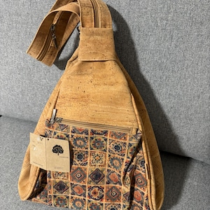 May include: A light brown cork sling bag with a patterned front pocket. The bag has a long adjustable strap and a zipper closure. The front pocket features a colorful geometric design with blue, orange, and brown squares. A small tag is attached to the bag.