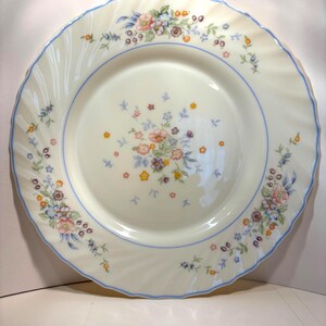 May include: Cream-colored dinner plate with a blue rim and floral design. The plate features a delicate pattern of pink, blue, yellow, and purple flowers. The edge has a wavy design.