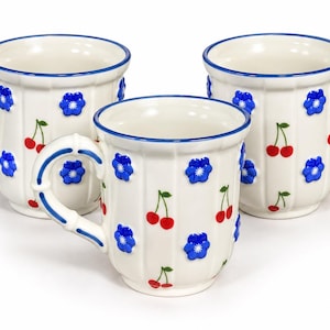 May include: Three white ceramic mugs with a ribbed texture, featuring raised blue flower appliqués and painted red cherry motifs. Each mug has a blue-rimmed edge and a handle decorated with blue stripes, creating a consistent set of drinkware.