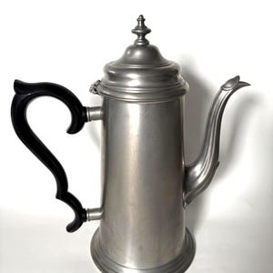 May include: A tall, metallic silver-toned coffee pot with a smooth, matte finish. It features a curved black handle, a long, slender spout, and a matching lid topped with a small, decorative finial. The base is circular and slightly flared.