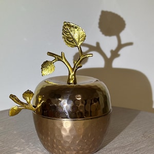 May include: A decorative apple-shaped container with a lid, crafted from gold-toned metal. The container features a textured, hammered finish on the lower half and a smooth, shiny top. A gold-colored branch with leaves serves as a handle and decorative element.