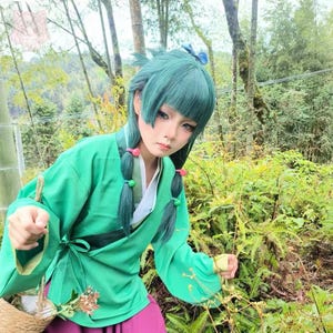May include: A person wearing a green cosplay outfit with a matching wig and braided pigtails. The outfit includes a green top with a tie closure and a white inner layer, paired with a purple skirt. The background features lush greenery.